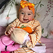 Isabella Witt joined the competition — help win amazing prizes! baby, child, smiling, headband, orange, striped_clothing, pumpkin, toy, plush, pillow, blanket, floral, cute, infant, happy, indoor, person, soft_toy, lying_down, cozy