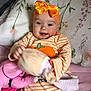 baby, child, smiling, headband, orange, striped_clothing, pumpkin, toy, plush, pillow, blanket, floral, cute, infant, happy, indoor, person, soft_toy, lying_down, cozy