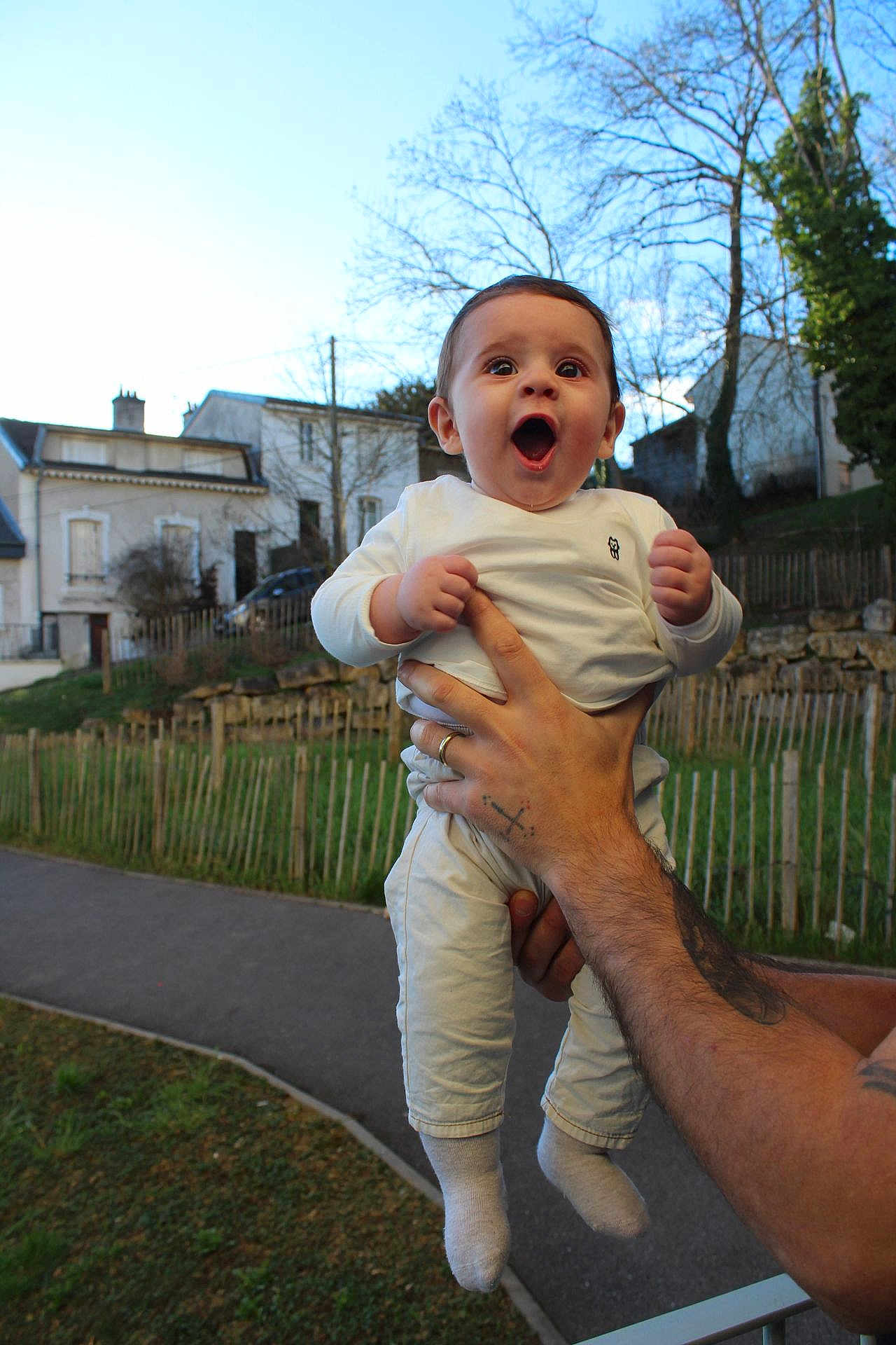 Vardan participe au concours pour gagner de l'argent avec cette photo : baby, infant, child, smiling, surprised, adult_hand, tattoo, wedding_ring, white_clothing, socks, outdoors, house, fence, grass, pathway, trees, sky, portrait, happy, holding