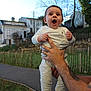 baby, infant, child, smiling, surprised, adult_hand, tattoo, wedding_ring, white_clothing, socks, outdoors, house, fence, grass, pathway, trees, sky, portrait, happy, holding