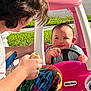 Violet is registered to the contest to win money with this photo: baby, toy_car, pink, child, smiling, steering_wheel, adult, food, sandwich, grass, outdoor, daylight, happy, person, play, colorful_pants, lawn, sidewalk, plastic, fun