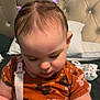 baby, child, infant, toddler, hair_ties, purple, onesie, orange, floral_pattern, bed, pillow, headboard, face, skin, arms, indoor, focused, cute, sitting, plastic_container