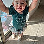 Violet is registered to the contest to win money with this photo: baby, bow, child, girl, glass_door, green_clothing, happy, headband, holding_hand, indoor, person, portrait, shadow, skirt, smiling, standing, sunlight, tile_floor, white_shoes, window
