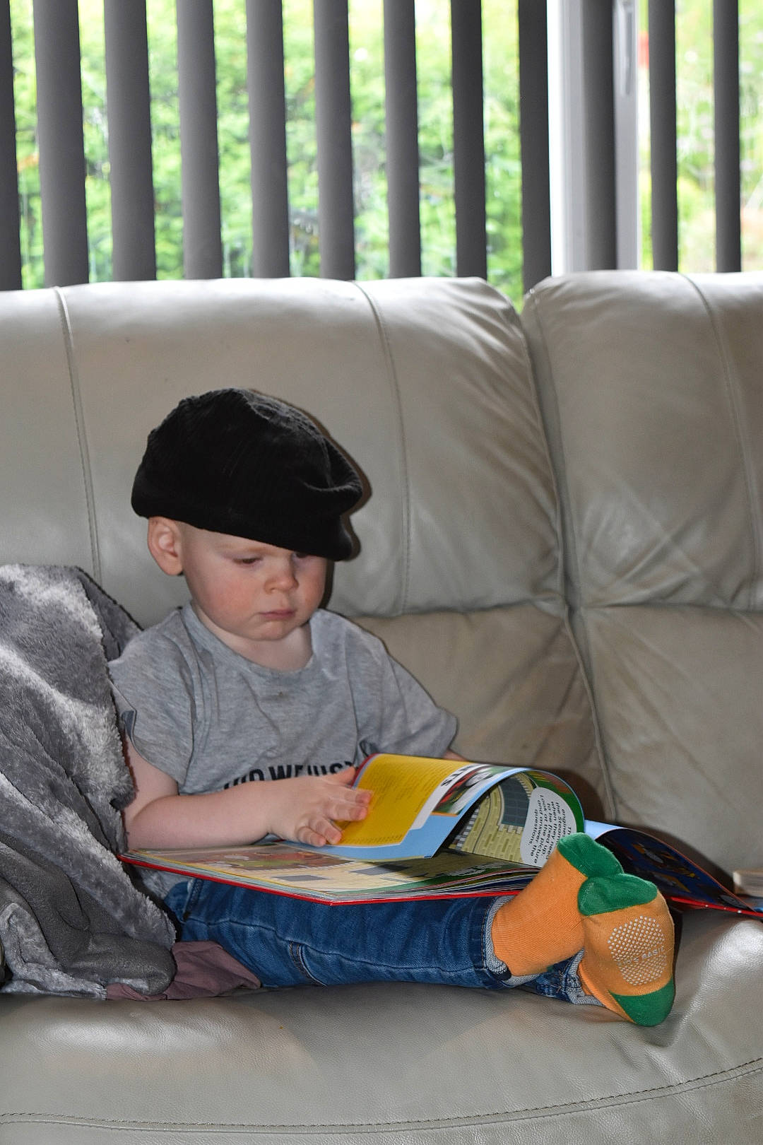 Jaxson joined the competition — help win amazing prizes! baseball_cap, book, cap, child, comfort, couch, flooring, hat, headwear, lap, leisure, living_room, nap, person, reading, room, shade, sitting, sleeve, t_shirt