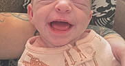 River joined the competition — help win amazing prizes! baby, newborn, infant, child, smiling, happy, portrait, close_up, face, eyes_closed, mouth_open, pink_outfit, giraffe_pattern, onesie, sitting, supported, adult, tattoo, indoor, cozy