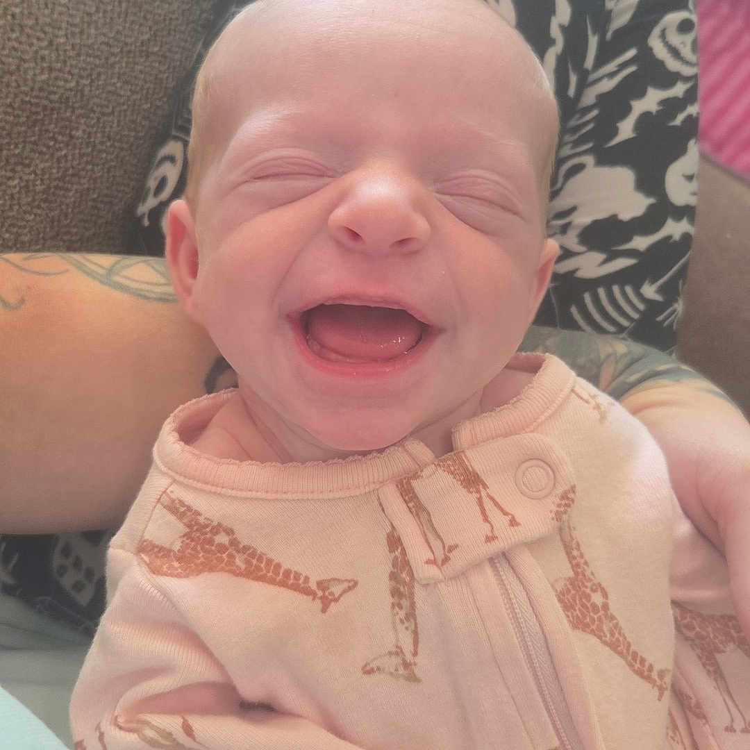 River joined the competition — help win amazing prizes! baby, newborn, infant, child, smiling, happy, portrait, close_up, face, eyes_closed, mouth_open, pink_outfit, giraffe_pattern, onesie, sitting, supported, adult, tattoo, indoor, cozy
