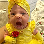 Andrea is registered to the contest to win money with this photo: baby, child, yellow_dress, headband, flower, rose, gloves, portrait, cute, infant, expression, hand, holding, indoor, fabric, person, face, bright, soft, adorable