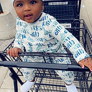 Quanzell is registered to the contest to win money with this photo: baby, child, shopping_cart, hat, pajamas, dinosaur_pattern, outdoor, sidewalk, curious, joyful, sparkles, metal_cart, clothing, footwear, person, infant, cute, playful, shopping, urban