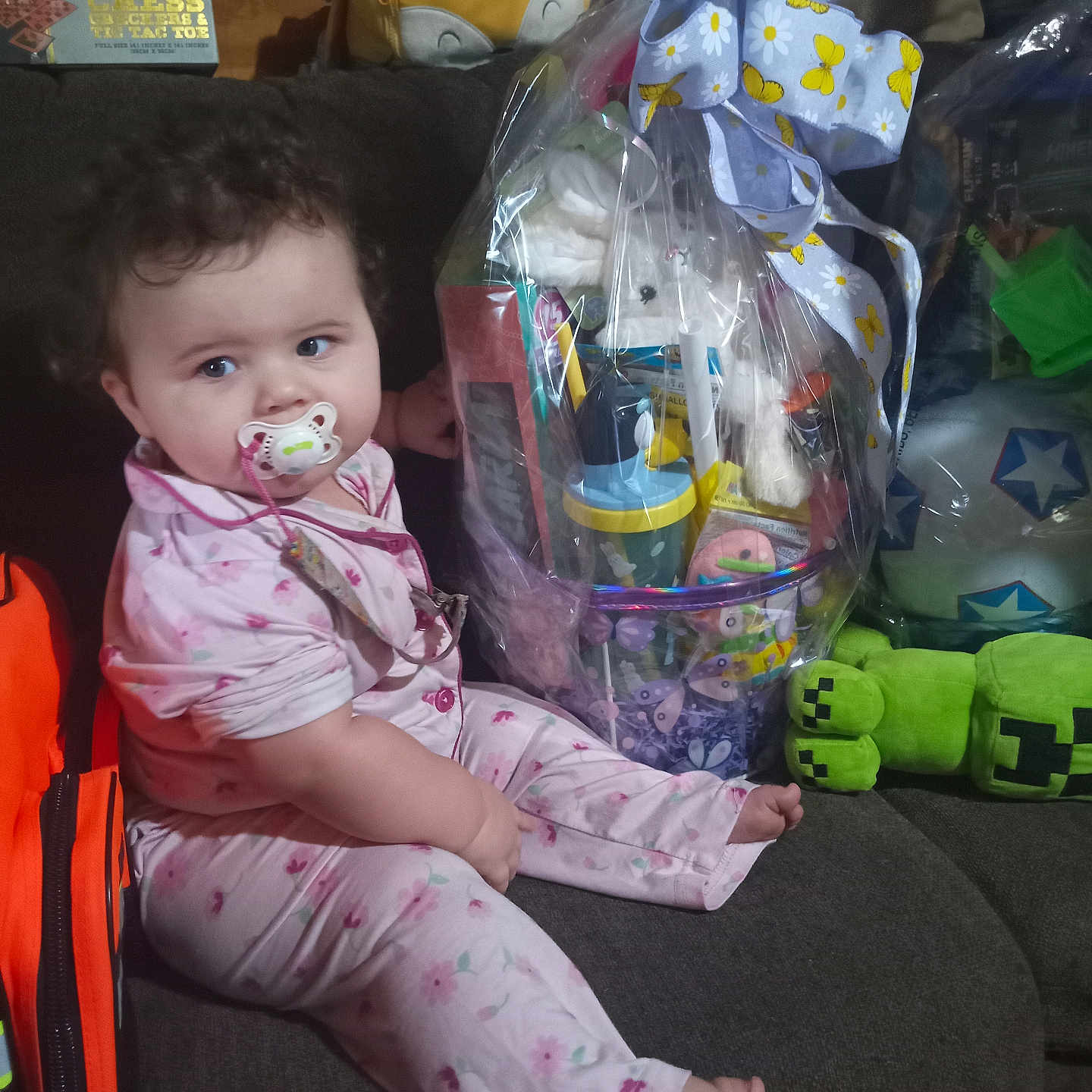 Heavenly is registered to the contest to win money with this photo: baby, pacifier, pajamas, couch, gift_basket, bow, toys, stuffed_animal, plastic_wrap, child, indoor, curly_hair, sitting, colorful, plush_toy, face, person, relaxing, home, comfort