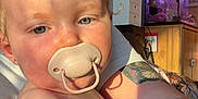 Daisy-Mae is registered to the contest to win money with this photo: baby, pacifier, face, blue_eyes, skin, arm, tattoo, sunlight, shadow, indoor, fish_tank, aquarium, cabinet, furniture, person, child, closeup, expression, home, relax