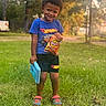 black_shorts, blue_shirt, boy, casual_clothing, child, chips, grass, happy, hot_wheels, lay's, nature, outdoor, slippers, smiling, snack, spiderman, standing, sunlight, toy, trees