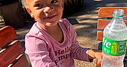 Arabella is registered to the contest to win money with this photo: child, smiling, outdoor, daylight, park, tree, bench, wooden_bench, pink_shirt, curly_hair, bottle, sparkling_water, table, casual_clothing, happy, nature, sunlight, person, young_child, portrait