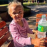 child, smiling, outdoor, daylight, park, tree, bench, wooden_bench, pink_shirt, curly_hair, bottle, sparkling_water, table, casual_clothing, happy, nature, sunlight, person, young_child, portrait