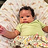 baby, infant, curly_hair, green_shirt, floral_skirt, blanket, crown_pattern, princess_theme, face, portrait, indoors, cute, child, person, skin, expression, resting, soft_texture, comfort, young