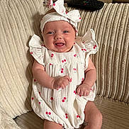 Emmie joined the competition — help win amazing prizes! baby, infant, smiling, headband, white_dress, cherry_pattern, couch, remote_control, happy, sitting, cute, indoors, soft_lighting, cozy, child, person, sock, furniture, home, portrait