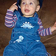 Maxieum joined the competition — help win amazing prizes! toddler, child, denim_overalls, dinosaur_patch, striped_shirt, cap, blue_cap, couch, brown_couch, indoor, sitting, curious, playful, footwear, socks, shoe, hand, face, person, young_child
