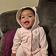 Malayah is registered to the contest to win money with this photo: baby, child, pink_onesie, hood, smiling, happy, sitting, blanket, couch, indoor, cute, infant, portrait, cozy, furniture, soft, clothing, young_child, person, warm