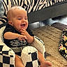 baby, child, infant, sitting, smiling, laughing, portrait, face, indoor, living_room, floor, rug, couch, zebra_pattern, blanket, checkered_pattern, clothing, black_shirt, baby_seat, happy