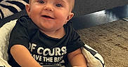 Gilberto is registered to the contest to win money with this photo: baby, child, infant, smiling, sitting, portrait, indoors, living_room, blanket, zebra_pattern, checkered_pattern, black_shirt, clothing, couch, furniture, carpet, happy, face, hands, feet