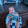 baby, infant, child, smiling, sitting, indoor, living_room, floor, blanket, checkered, baby_seat, safety_harness, blue_shirt, bare_feet, television, stroller, sofa, portrait, happy, family_home