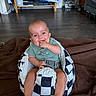 baby, child, infant, smiling, sitting, indoor, living_room, portrait, face, hands, fingers, green_shirt, blanket, patterned_seat, seat_belt, couch, television, stroller, wood_floor, happy