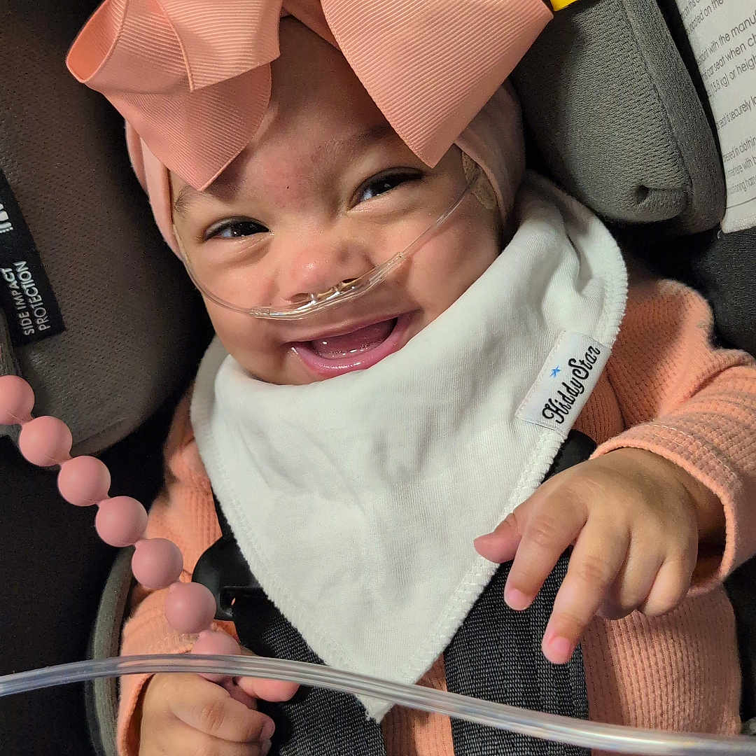 Sirene is registered to the contest to win money with this photo: accessory, baby, bib, car_seat, child, closeup, clothing, cute, face, hand, happy, headwear, indoor, infant, medical, nasal_cannula, pink_bow, seatbelt, smiling, toy