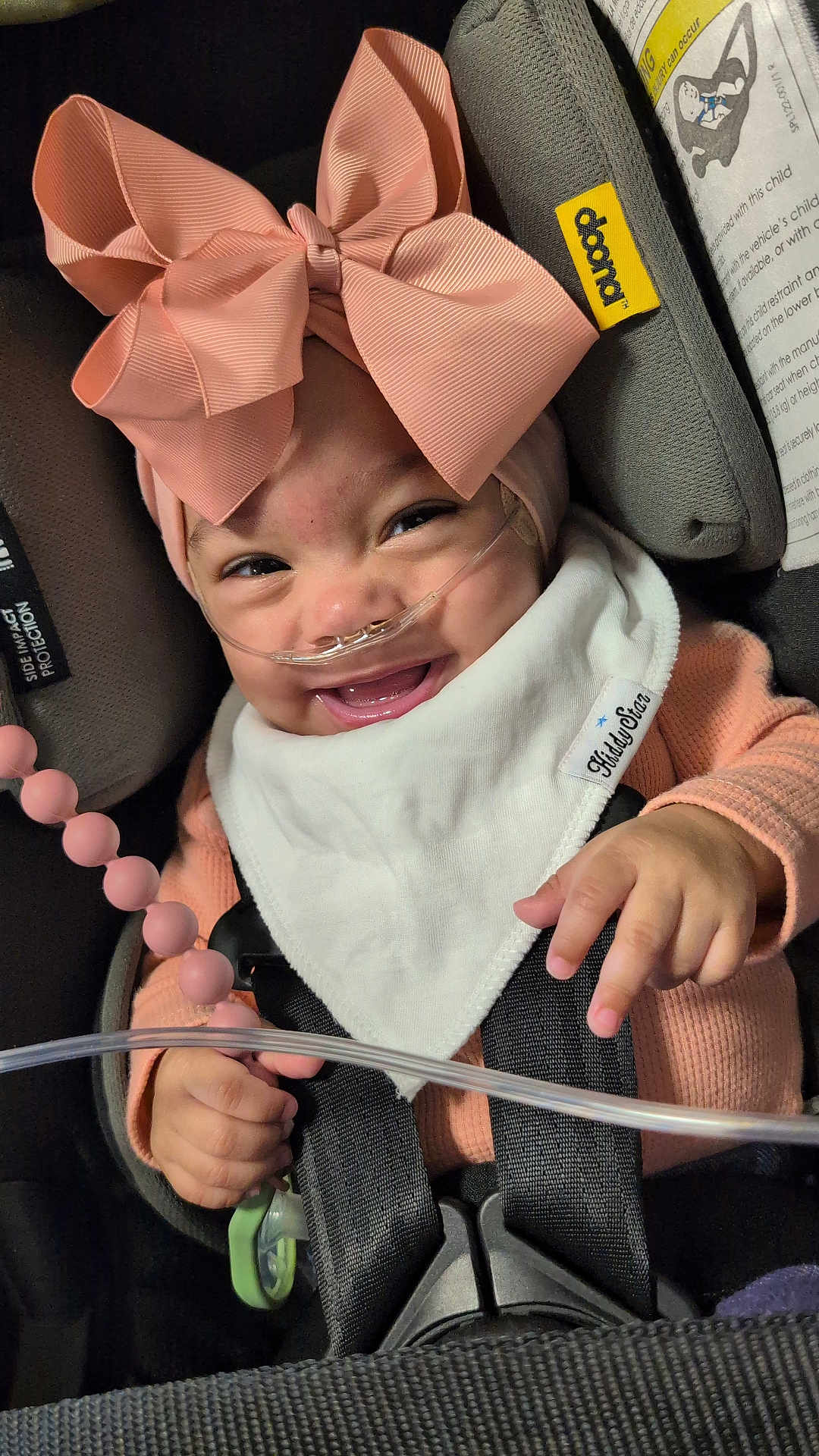 Sirene is registered to the contest to win money with this photo: baby, smiling, pink_bow, car_seat, nasal_cannula, toy, bib, child, infant, cute, happy, seatbelt, clothing, hand, face, headwear, indoor, closeup, medical, accessory