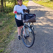Ullis a rejoint le concours — aidez-le/la à gagner de superbes lots ! bicycle, bike_basket, casual_clothing, cycling, dog, forest, glasses, greenery, helmet, leisure, nature, outdoor, path, person, pet, road, shorts, sunlight, trees, white_bicycle