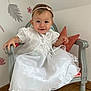 baby, child, girl, white_dress, headband, smile, chair, cushion, floor, wooden_floor, wall, flower_decor, leaf_decor, indoors, portrait, cute, sitting, happy, person, footwear