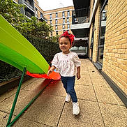 Sophia joined the competition — help win amazing prizes! child, toddler, girl, red_bow, white_shirt, blue_jeans, white_sneakers, slide, playground, urban, courtyard, brick_building, pavement, greenery, outdoor, daylight, smiling, walking, happy, play