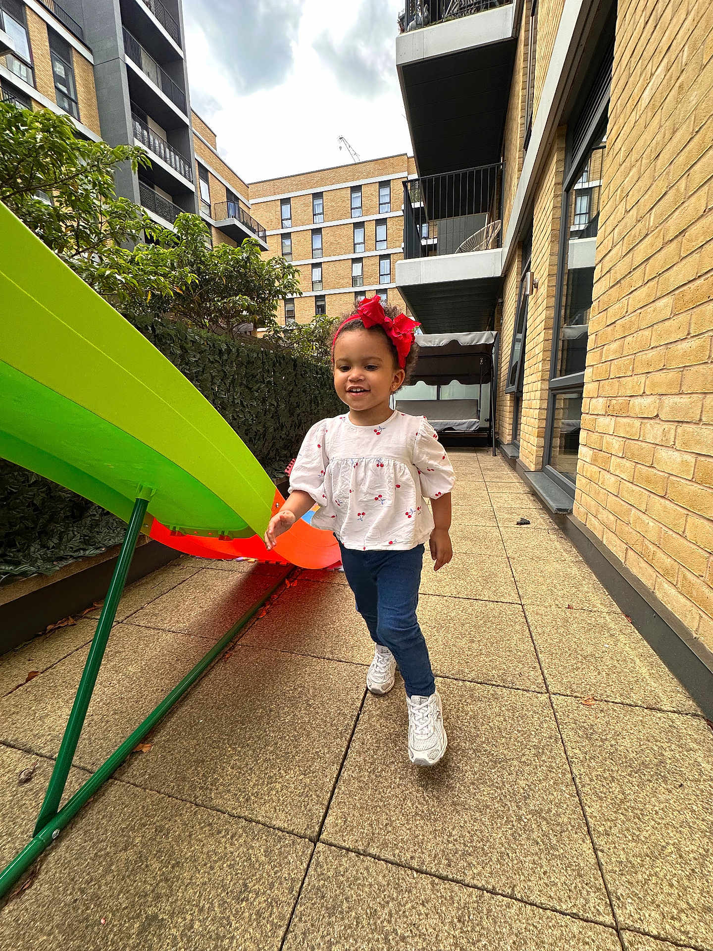 Sophia joined the competition — help win amazing prizes! child, toddler, girl, red_bow, white_shirt, blue_jeans, white_sneakers, slide, playground, urban, courtyard, brick_building, pavement, greenery, outdoor, daylight, smiling, walking, happy, play
