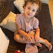 Aaliyah a rejoint le concours — aidez-le/la à gagner de superbes lots ! accessories, barefoot, child, colorful, couch, cross_legged, curly_hair, cute, dress, floral_pattern, girl, happy, home, indoor, living_room, person, pillow, playful, smile, toy