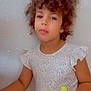child, curly_hair, dress, patterned_clothing, toy, indoor, standing, holding, person, young_child, casual, neutral_expression, wall, light_background, short_sleeves, colorful, small_toy, portrait, single_person, cute