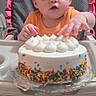 baby, child, high_chair, cake, sprinkles, orange_shirt, indoor, birthday_cake, food, celebration, curious, small_hands, pink_ruffle, face, person, table, home, cute, sitting, young_child