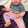toddler, child, baby, sitting, blue_eyes, colorful_clothing, tie_dye, tshirt, indoor, blanket, plush, foot, hand, face, curly_hair, bedroom, soft_texture, casual, portrait, cute