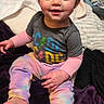 Athena is registered to the contest to win money with this photo: toddler, child, sitting, colorful_clothing, tie-dye, graphic_tee, barefoot, indoor, blanket, purple, smiling, curly_hair, bedroom, toys, box, fabric, cozy, happy, person, cute