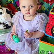 Athena joined the competition — help win amazing prizes! baby, child, girl, lavender_dress, headband, blue_eyes, toy, lollipop, colorful_blanket, floral_pattern, plush_toy, couch, indoor, cute, sitting, portrait, playtime, soft_lighting, expression, infant