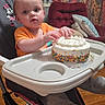 toddler, high_chair, cake, orange_shirt, sprinkles, hands, wooden_floor, curtains, cushions, living_room, footwear, baby, table, face, person, indoor, furniture, celebration, birthday_cake, expression