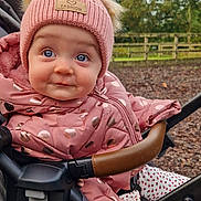 Eibhlin joined the competition — help win amazing prizes! baby, child, stroller, pink_jacket, knit_hat, blue_eyes, outdoor, fall_leaves, cute, drool, polka_dots, warm_clothing, infant, bundle_up, face, expression, person, nature, trees, fence