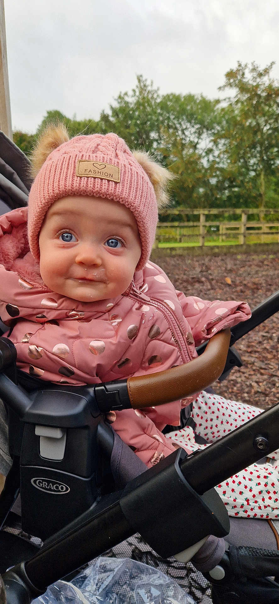 Eibhlin joined the competition — help win amazing prizes! baby, child, stroller, pink_jacket, knit_hat, blue_eyes, outdoor, fall_leaves, cute, drool, polka_dots, warm_clothing, infant, bundle_up, face, expression, person, nature, trees, fence