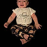 baby, child, smiling, sitting, white_shirt, patterned_pants, cowboy_hat_graphic, text_on_shirt, happy, cute, infant, portrait, isolated_background, black_background, clothing, person, studio_style, young_child, feet, hands