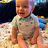baby, child, smiling, sitting, overalls, blue_shirt, floral_quilt, indoor, face, happy, cute, infant, furniture, blanket, household_items, toys, skin, foot, hand, portrait
