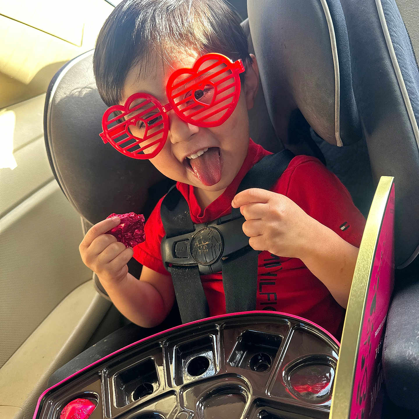 Jonathan is registered to the contest to win money with this photo: child, car_seat, red_sunglasses, heart_shaped_glasses, tongue_out, red_shirt, chocolate, chocolate_box, seat_belt, car_interior, playful, happy, smiling, person, kid, fun, snack, seat, window, sunlight