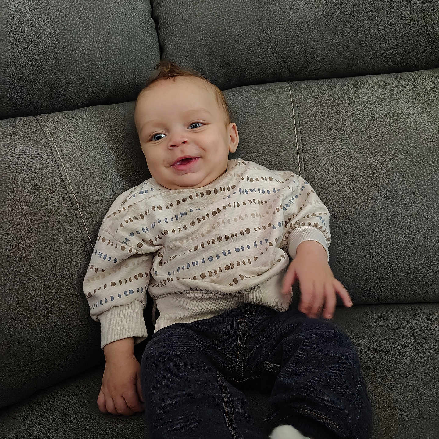 Asher is registered to the contest to win money with this photo: baby, child, infant, smiling, happy, portrait, sitting, couch, sofa, furniture, indoor, home, sweater, clothing, jeans, socks, face, eyes, brown_hair, cozy