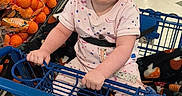Luna is registered to the contest to win money with this photo: baby, shopping_cart, grocery_store, produce, oranges, child, infant, floor, clothing, socks, person, supermarket, blue, holding, seated, aisle, food, basket, retail, cute
