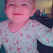 Luna is registered to the contest to win money with this photo: baby, child, smiling, floral_clothing, onesie, indoor, face, happy, sparkle_filter, cute, portrait, infant, young_child, reaching, joyful, cozy, home, person, pink, soft_lighting