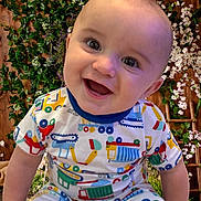 Kaleb joined the competition — help win amazing prizes! baby, child, smiling, face, pajamas, colorful_clothing, sitting, indoor, wooden_wall, flower, greenery, happy, cute, portrait, infant, playful, bright_colors, person, closeup, joyful