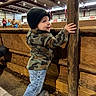 barn, beanie, boy, camouflage_jacket, child, equestrian, face, horse, horseback_riding, indoor, jeans, metal_post, portrait, reaching, riding_arena, smiling, standing, toddler, winter_clothing, wooden_fence