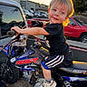 child, toddler, boy, smiling, portrait, dirt_bike, motorcycle, motocross, outdoor, sunset, golden_hour, vehicle, handlebars, helmetless, black_shirt, shorts, sneakers, parked_cars, street, urban_background