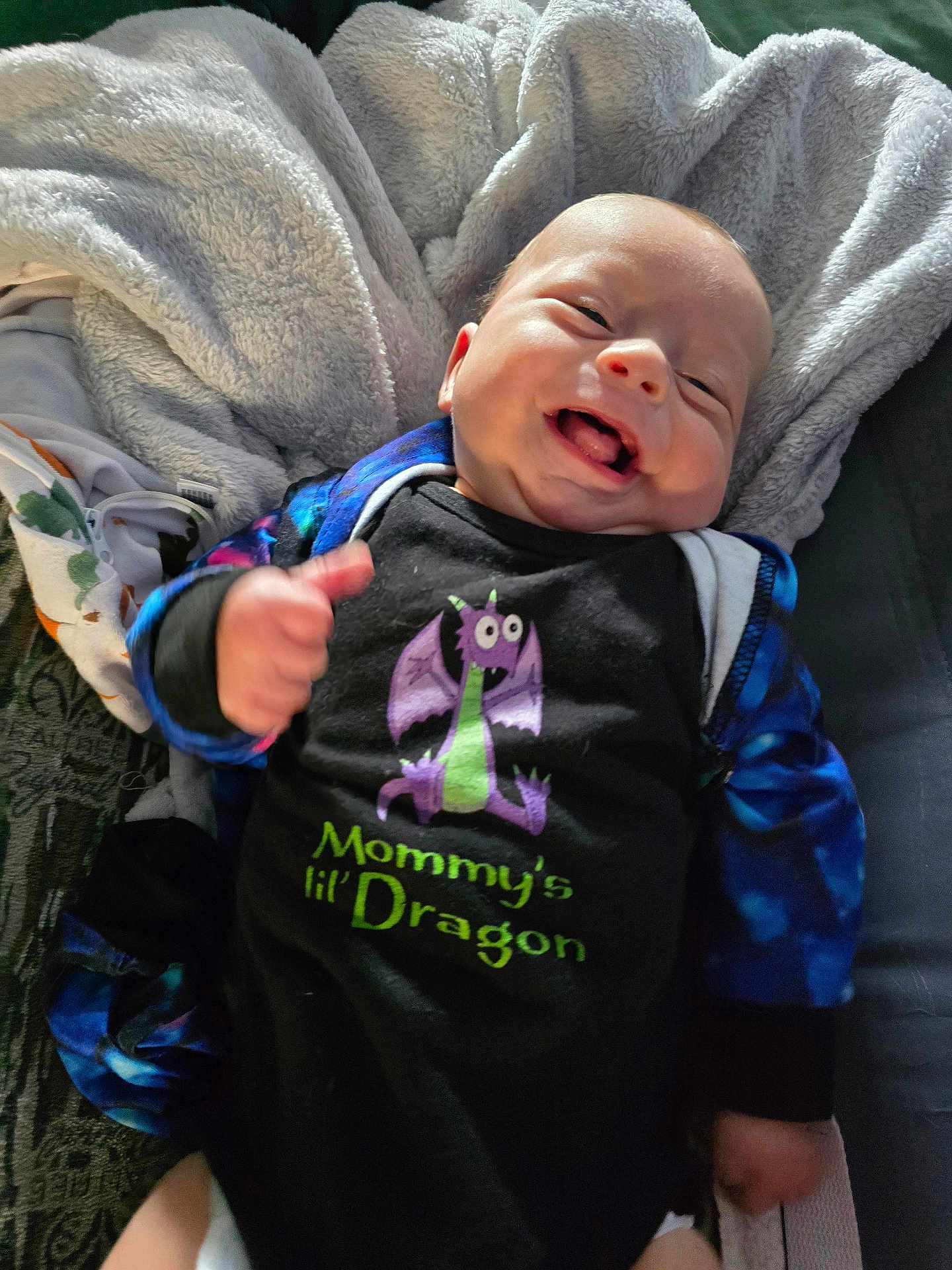 Leon joined the competition — help win amazing prizes! baby, smiling, onesie, dragon, text_on_clothing, blanket, happy, infant, thumbs_up, cute, child, clothing, portrait, indoors, person, fabric, face, sleeves, sitting, young_child